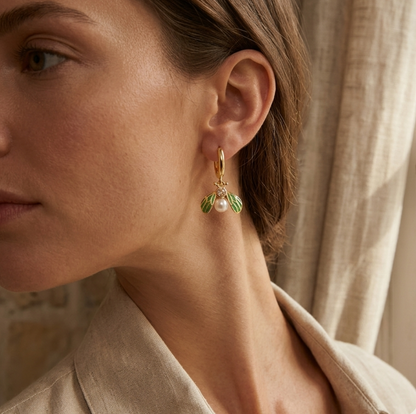 Close-up of a woman wearing gold earrings with green leaves and a pearl in front of a beige curtain.
