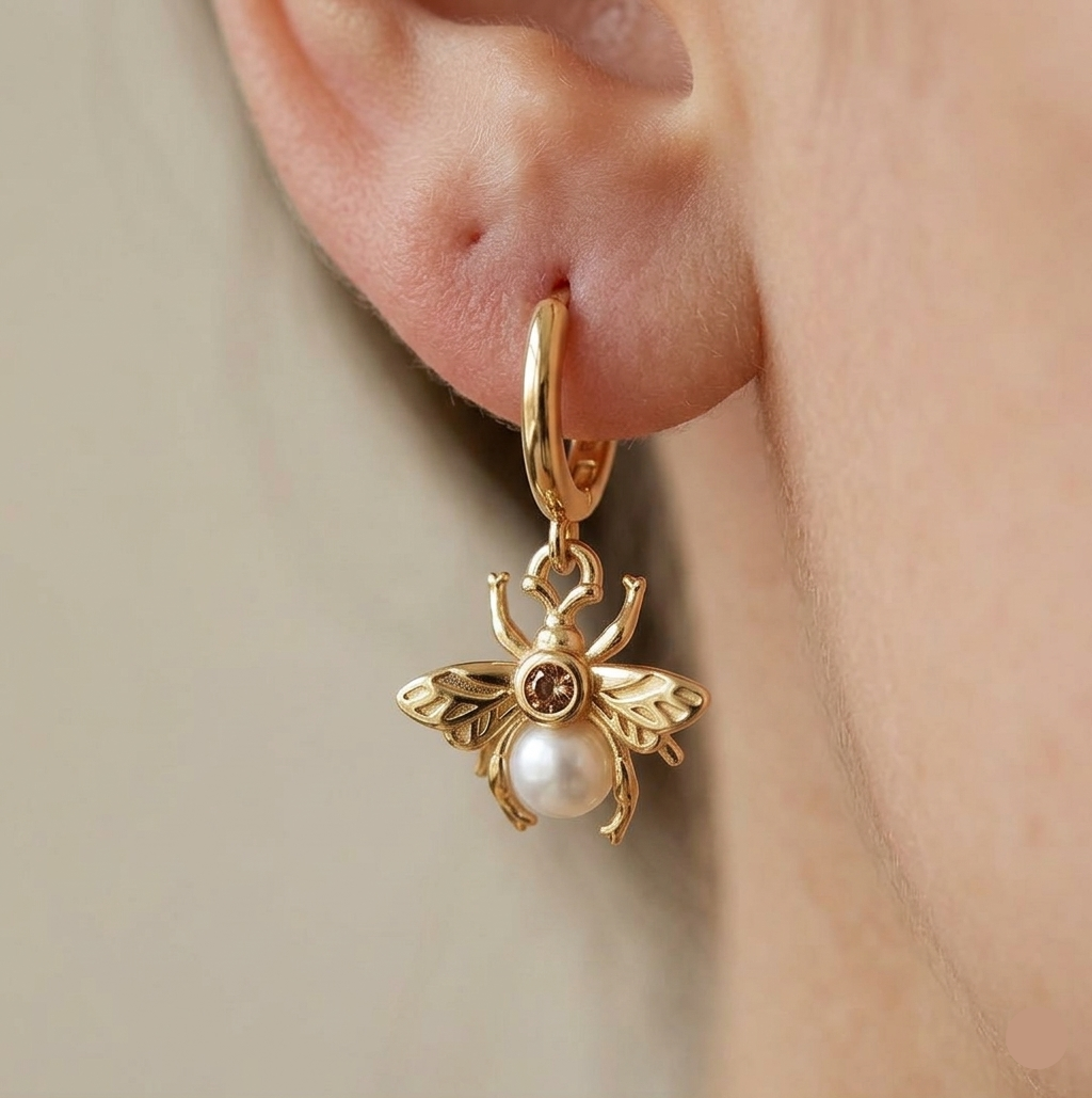 Gold bee-shaped earring with a pearl on an ear against a neutral background