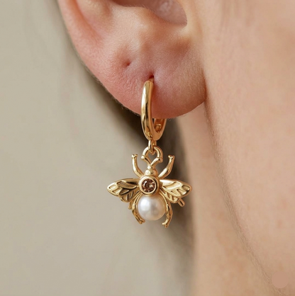 Gold bee-shaped earring with a pearl on an ear against a neutral background